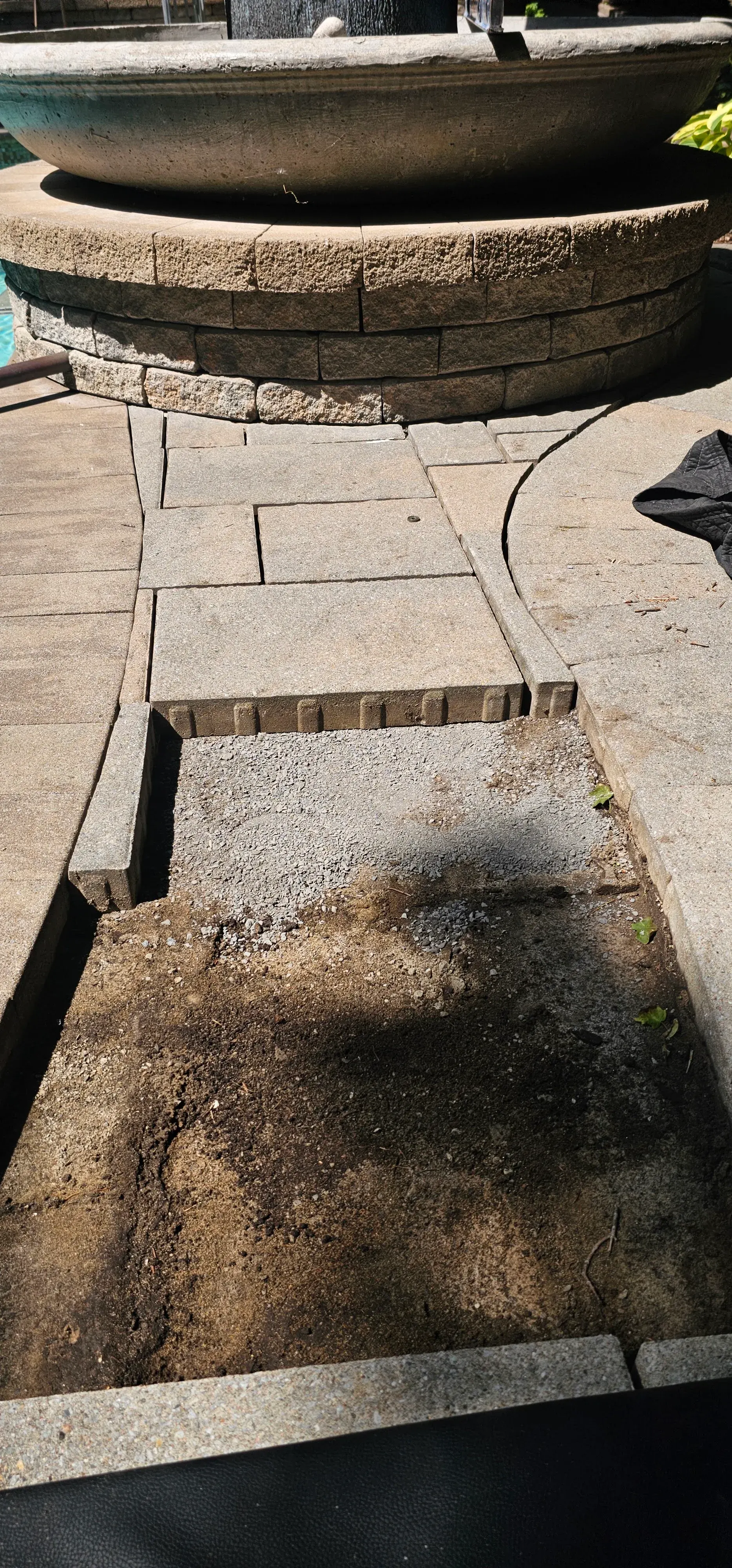 Pavers lifted beside a fountain ring, exposing the gravel bedding, soil and edge restraint beneath the surface