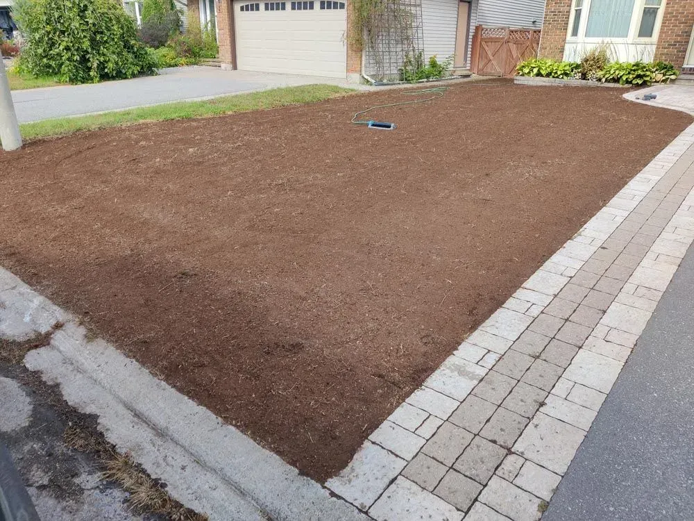 Front lawn stripped to a level, prepared seedbed of dark soil, paver border down the right side to the street curb