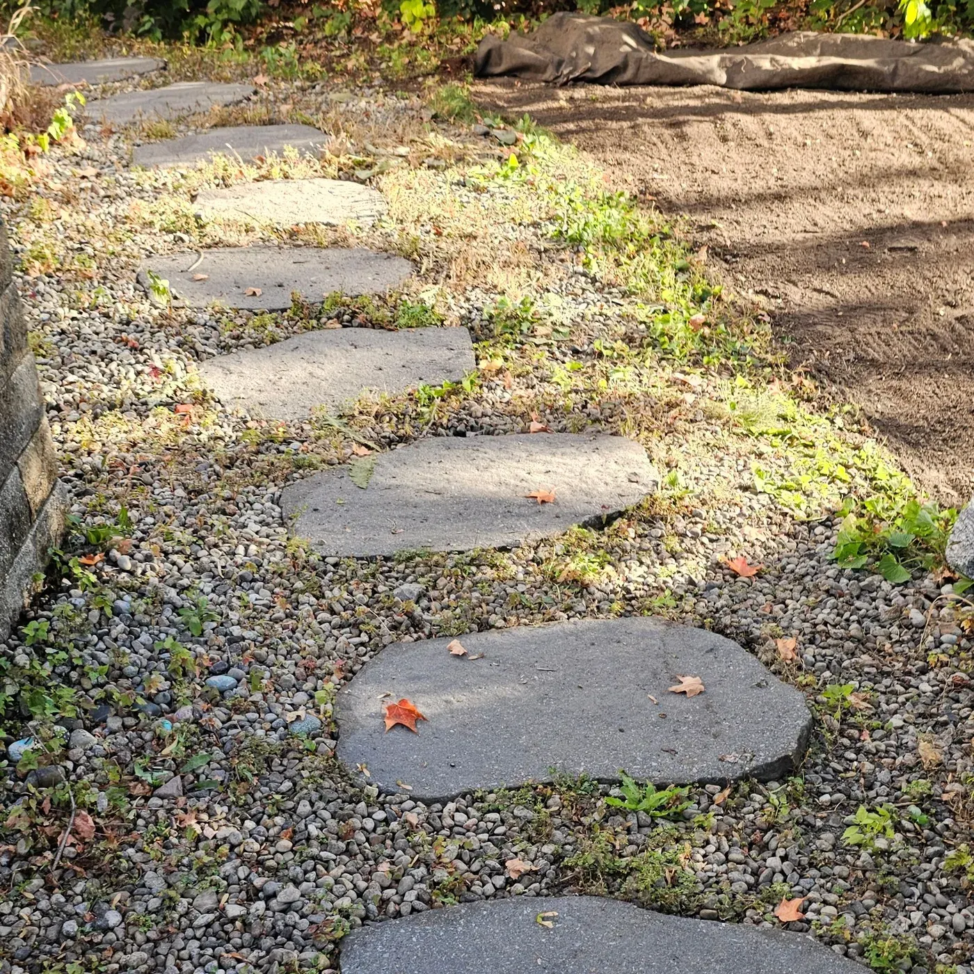 A river rock path with large flagstone stepping stones beside a stone pillar, grass and weeds growing through the stone around every step